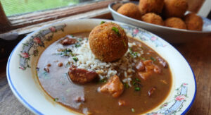 bowl of gumbo topped with a potato salad croquette, with a platter of croquettes in background