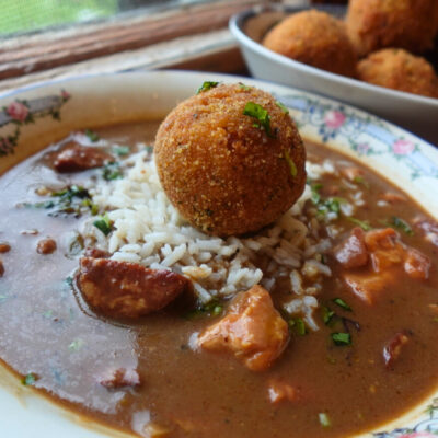 bowl of gumbo topped with a potato salad croquette, with a platter of croquettes in background