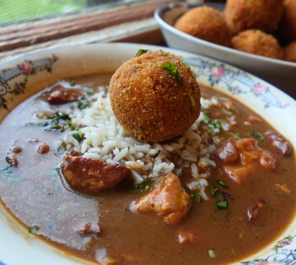 bowl of gumbo topped with a potato salad croquette, with a platter of croquettes in background